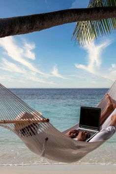 a man laying in a hammock on the beach using a laptop computer while relaxing