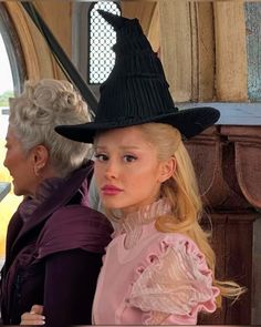 an older woman wearing a witches hat and looking at another woman in a pink dress