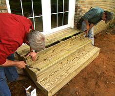 two men working on a wooden bench in front of a house