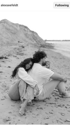 a man and woman sitting on the beach with their arms around each other, hugging