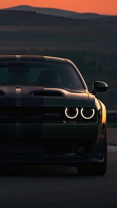 the front end of a black and white car with its lights turned on at sunset