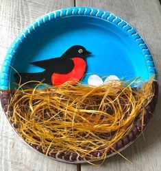 a paper plate with a bird and eggs in it on a wooden table next to straw