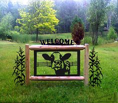 a welcome sign in the middle of a grassy field with trees and bushes behind it