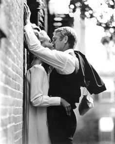 a man and woman leaning against a brick wall, one kissing the other's forehead