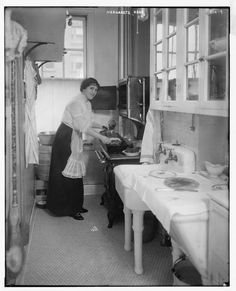 an old black and white photo of a woman cooking in a kitchen with two sinks