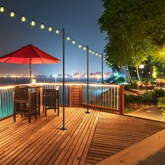 an outdoor deck with chairs and umbrellas overlooking the water at night, lit up by string lights