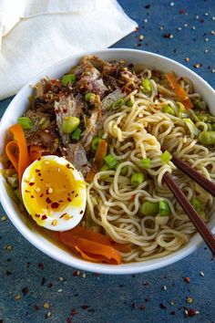 a white bowl filled with noodles, meat and veggies next to an egg