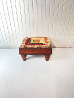 a small wooden bench with a patchwork seat cushion on the top and bottom, in front of a white wall