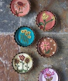 four bottle caps with flowers painted on them sitting on a stone floor next to a brick wall