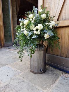 a large metal vase filled with white flowers sitting on top of a stone floor next to a wooden door