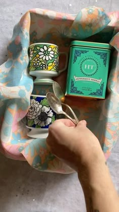 a person holding a spoon in front of a cup and tea tin with the lid open