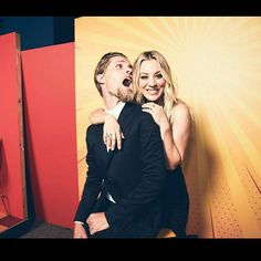 a man and woman standing next to each other in front of a red wall with an advertisement behind them