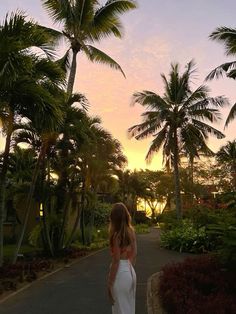 a woman in a white dress is walking down a path with palm trees behind her