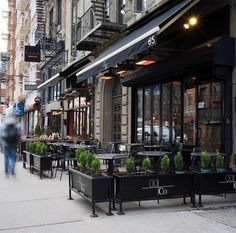 people walking down the sidewalk in front of an outdoor restaurant with tables and chairs on it