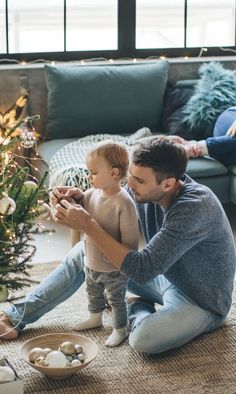 a man sitting on the floor holding a baby next to a christmas tree and other decorations