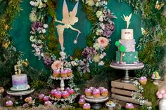 a table topped with cakes and cupcakes on top of a green wall covered in greenery