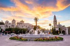 a fountain in the middle of a plaza with palm trees and buildings behind it at sunset