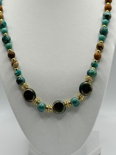 The beads are predominantly in shades of turquoise and brown with a glossy finish, and some have a marbled effect that adds depth and interest to their appearance. Between the larger beads are ornate gold-toned spacer beads with intricate designs, contributing to the necklace's luxurious look. The combination of the earthy brown tones with the vibrant turquoise creates a visually striking contrast. This necklaceswould appeal to those with a taste for bold, statement jewelry that combines natural Gold Turquoise Necklace With Round Gemstone Beads, Gold Turquoise Necklace With Round Beads And Natural Stones, Turquoise Malachite Necklaces With Round Beads, Gold Agate Necklace With Colorful Beads, Gold Jade Beaded Necklaces With Round Beads, Gold Turquoise Necklace With Round Beaded Details, Gold Jade Round Beaded Necklaces, Elegant Turquoise Agate Beaded Necklaces, Turquoise Agate Round Beads Gems And Cabochons