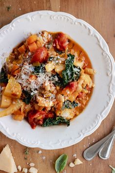 a white bowl filled with pasta and vegetables on top of a wooden table next to silverware