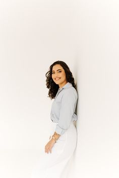 a woman leaning against a white wall with her hands on her hips and smiling at the camera