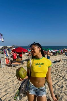 a woman in yellow shirt holding a coconut and watermelon on beach next to umbrellas