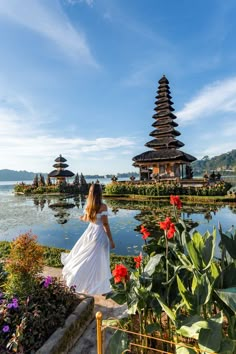 a woman in white dress walking by water with flowers and pagodas on the other side