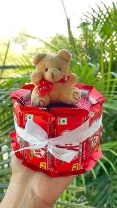 a person holding up a small teddy bear in a red gift box with white ribbon