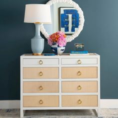 a white dresser topped with a blue vase and pink flowers next to a round mirror