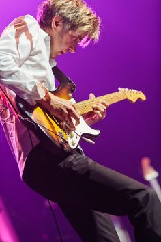 a man playing a guitar on stage with purple lighting behind him and his legs in the air