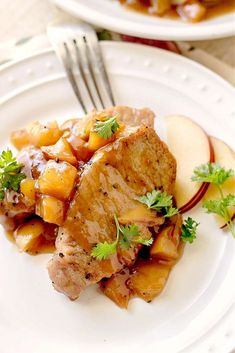 a white plate topped with meat and fruit covered in sauce next to a fork on top of a table