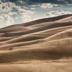 two people are walking in the desert with sand dunes and clouds above them, as seen from below