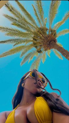 a woman in a yellow swimsuit and sunglasses standing under a palm tree with her eyes closed