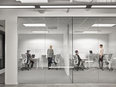 three people sitting at desks in an office with glass walls and white cubicles