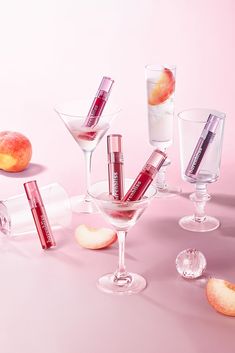 an assortment of lipstick and lip balm in glasses on a pink background with peaches