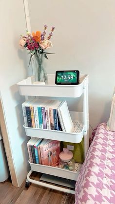 a white shelf with books on it next to a bed and flowers in a vase