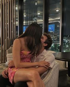 a man and woman sitting on a couch in front of a window with the city lights behind them