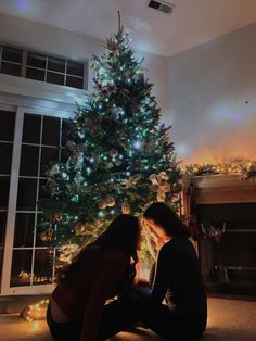 two people sitting in front of a christmas tree with lights on it and a lit fireplace