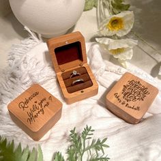 three wooden boxes with wedding rings inside on a white blanket next to flowers and an egg