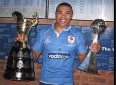 a man holding two trophies in front of a brick wall with plaques on the wall behind him