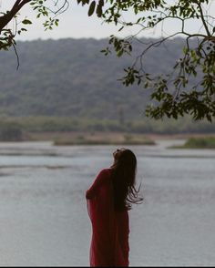 a woman in a red dress looking out over the water from behind a tree branch