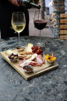 a person pouring wine into a glass next to some food on a cutting board with grapes, cheese and crackers