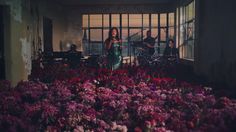 a woman standing in front of a window surrounded by purple and red flowers with two men playing drums behind her