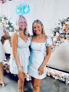 two women standing next to each other in front of a floral backdrop and neon sign