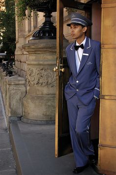 a man in a blue suit and bow tie leaning against a door with his hands on his hips