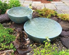 two water bowls sitting on top of rocks in a garden next to a small statue