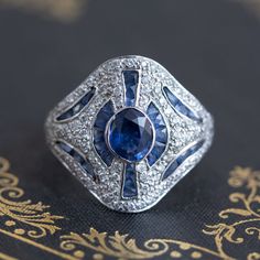 A fun and unusual cocktail ring, the styling of this ring is reminiscent of Art Deco themes with the geometric lines and the use of diamonds alongside blue sapphires. The central stone is a faceted medium blue oval sapphire of approximately 1.00ct, surrounded by 28 calibre cut sapphires (approx. .75ctw); there are also .50ctw of diamonds added in for good measure to yield an ornate yet subtly fancy ring! The setting is low-profile making it very easy and comfortable to wear. 18kt white gold; siz Art Deco Blue Diamond Ring With Center Stone, Art Deco Blue Sapphire Ring With Center Stone, Blue Art Deco Diamond Ring With Center Stone, Art Deco Blue Diamond Ring, Blue Diamond Art Deco Ring, Blue Art Deco Diamond Ring, Art Deco Blue Diamond Ring With Accents, White Gold Sapphire Art Deco Ring, Blue Art Deco Diamond Ring With Diamond Cut