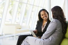 two women in business attire sitting on a couch and one is holding a clipboard