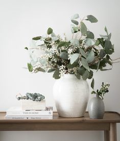 a white vase filled with lots of green leaves on top of a wooden table next to books