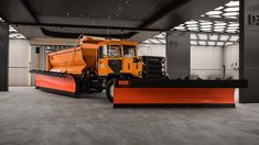 a large orange truck parked inside of a building next to a snow plow on the ground