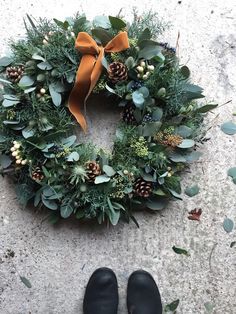 a wreath with pine cones and greenery on the ground next to a pair of black slippers